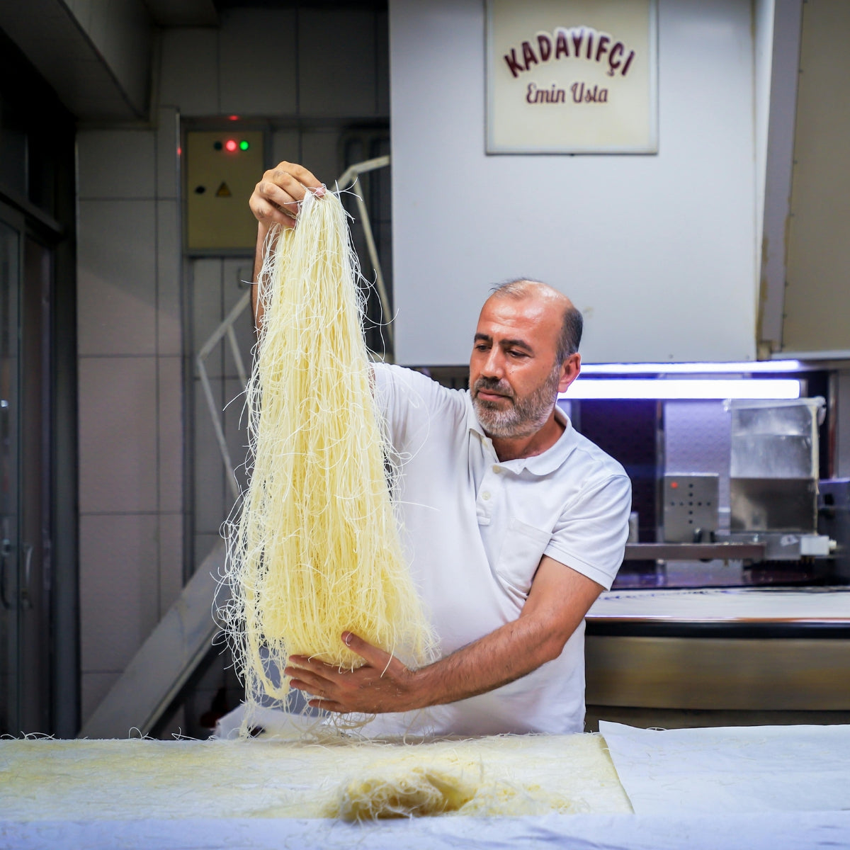 In a kitchen under a "Kadayıfçı Emin Usta" sign, a man in a white shirt carefully holds strands of Theos Fresh Kadayif (Kataifi) Shredded Fillo Dough, evoking the expertise found at a renowned Dubai Chocolate Bar. In a kitchen under a "Kadayıfçı Emin Usta" sign, a man in a white shirt carefully holds strands of Theos Fresh Kadayif (Kataifi) Shredded Fillo Dough, evoking the expertise found at a renowned Dubai Chocolate Bar.