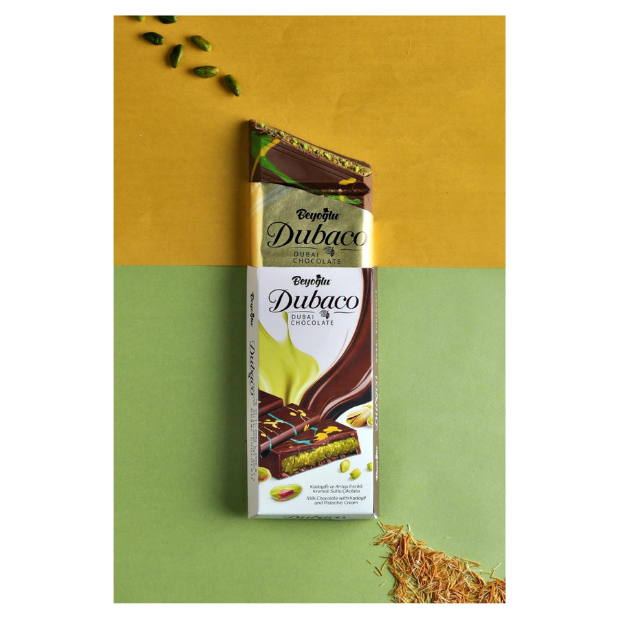 The "Beyoglu - Dubaco Premium Dubai Chocolate Bar - 200g" is partly unwrapped on a dynamic green and yellow background, surrounded by pistachios and spices, suggesting the exotic allure of Dubai Chocolate with Pistachio Cream.