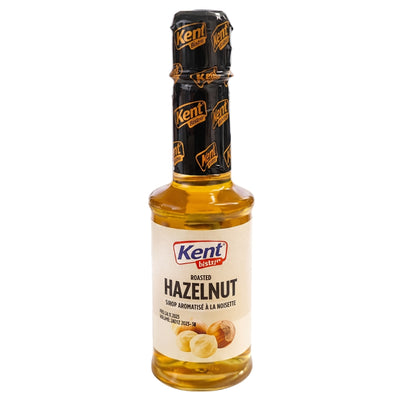 A bottle of Kent Roasted Hazelnut syrup, featuring a clear glass bottle filled with golden-brown liquid, a cream-colored label with hazelnut illustrations, and a black foil neck seal with gold branding.