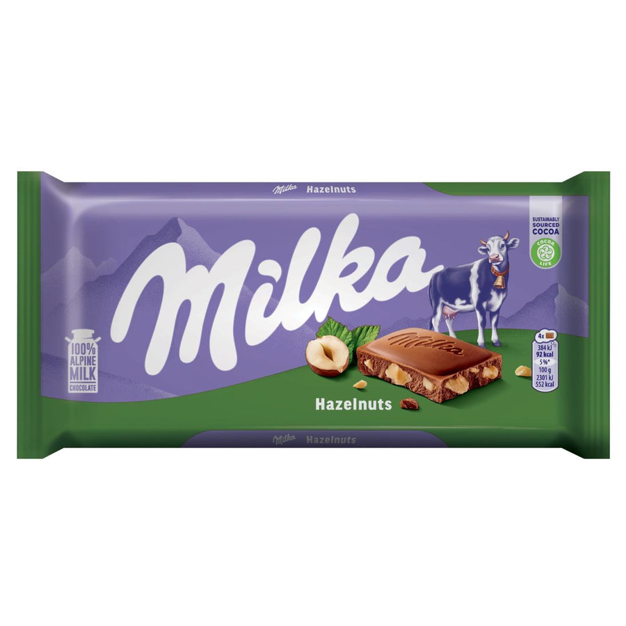 Milka - Chocolate With Hazelnut Pieces - 90g: Alpine milk chocolate with hazelnut pieces in signature purple Milka packaging, featuring a cow, mountains, a hazelnut, and the Milka logo.