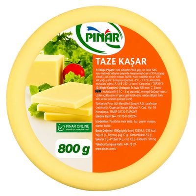 A round package of PİNİR Taze Kaşır cheese, showing the front label with product details, a photo of cheese wedges with herbs and a tomato, and an orange background on a white studio backdrop.