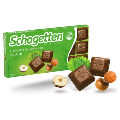 A sweet treat, the Schogetten Alpine Milk Chocolate Bar with Hazelnuts (100g) features individual chocolate squares, hazelnuts, and green leaves on its packaging.