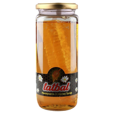 A clear glass jar labeled "Tatbal - Honeycomb in Honey Syrup Jar - 600g" contains honey syrup with a honeycomb piece inside. The label displays white flowers and gold accents, highlighting this pure, unprocessed Tatbal treat.
