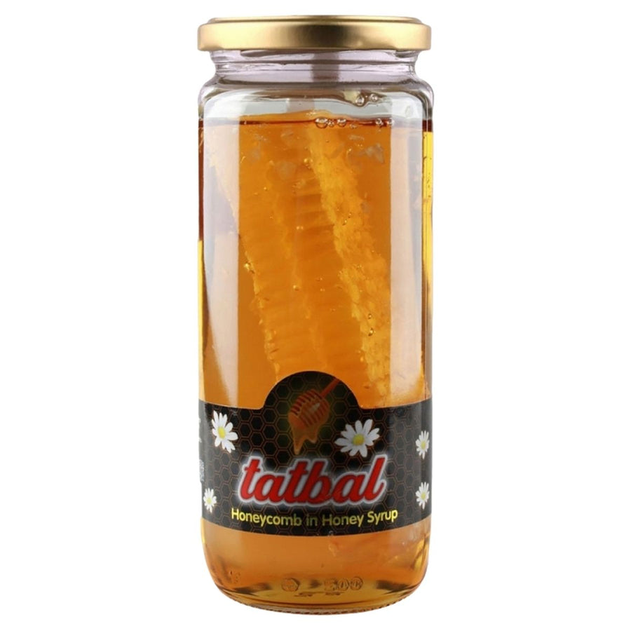 A clear glass jar labeled "Tatbal - Honeycomb in Honey Syrup Jar - 600g" contains honey syrup with a honeycomb piece inside. The label displays white flowers and gold accents, highlighting this pure, unprocessed Tatbal treat.