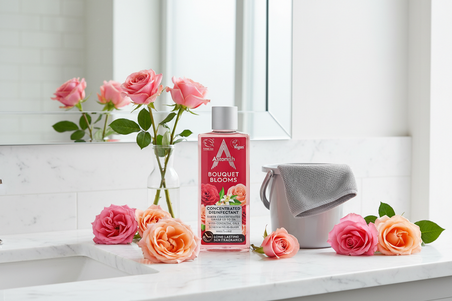 An Astonish Bouquet Blooms Concentrated Disinfectant (300ml), accompanied by a gray cleaning cloth and a small bucket, is arranged alongside fresh roses on a white bathroom countertop in front of a mirror.